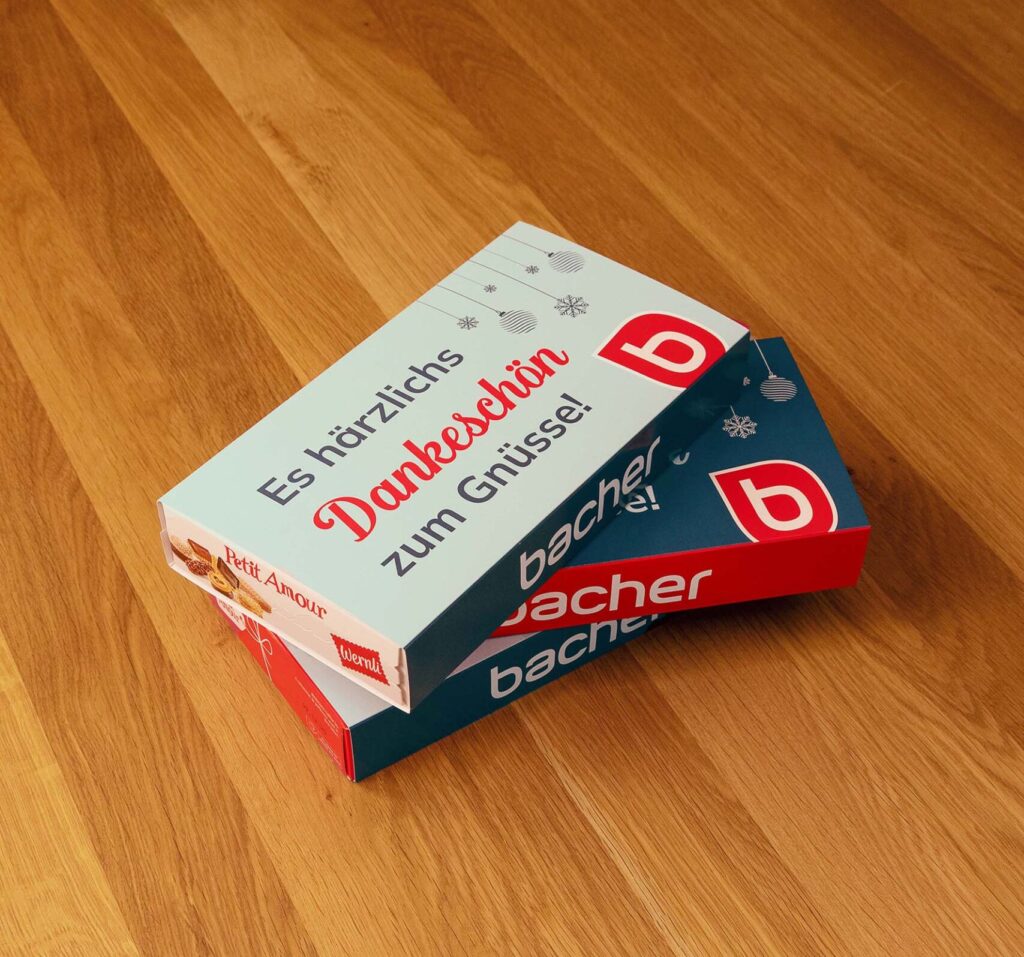 Eine blau-weiße Schachtel mit deutschem Text, darunter "Es herzliches Dankeschön zum Genießen!" und der Markenname "bacher", liegt leicht geöffnet auf einer Holzoberfläche - perfekt für ein Weihnachtsgeschenk.