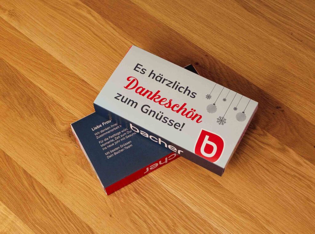 Eine Karte mit dem deutschen Text "Es herzlichs Dankeschön zum Gnuuss!" und einem roten "b"-Logo liegt auf einem Holztisch und verdeckt teilweise eine andere blaue und rote Karte darunter - perfekt für Weihnachtsgrüße.