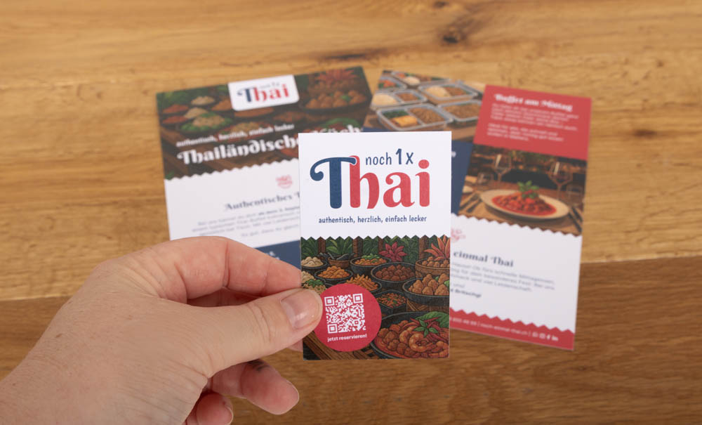 Eine Hand hält eine bunte Karte mit der Aufschrift "noch 1x Thai" über Broschüren mit Bildern von thailändischen Gerichten auf einem Holztisch.