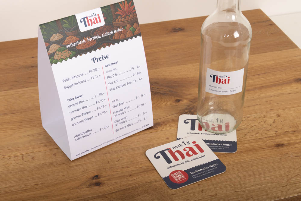 Auf einem Tisch liegen eine gefaltete Speisekarte eines thailändischen Restaurants, eine leere Glasflasche und zwei Markenuntersetzer auf einer Holzfläche. Auf den Gegenständen sind das Logo des Restaurants und die Menüpreise in Deutsch und Schweizer Franken angegeben.
