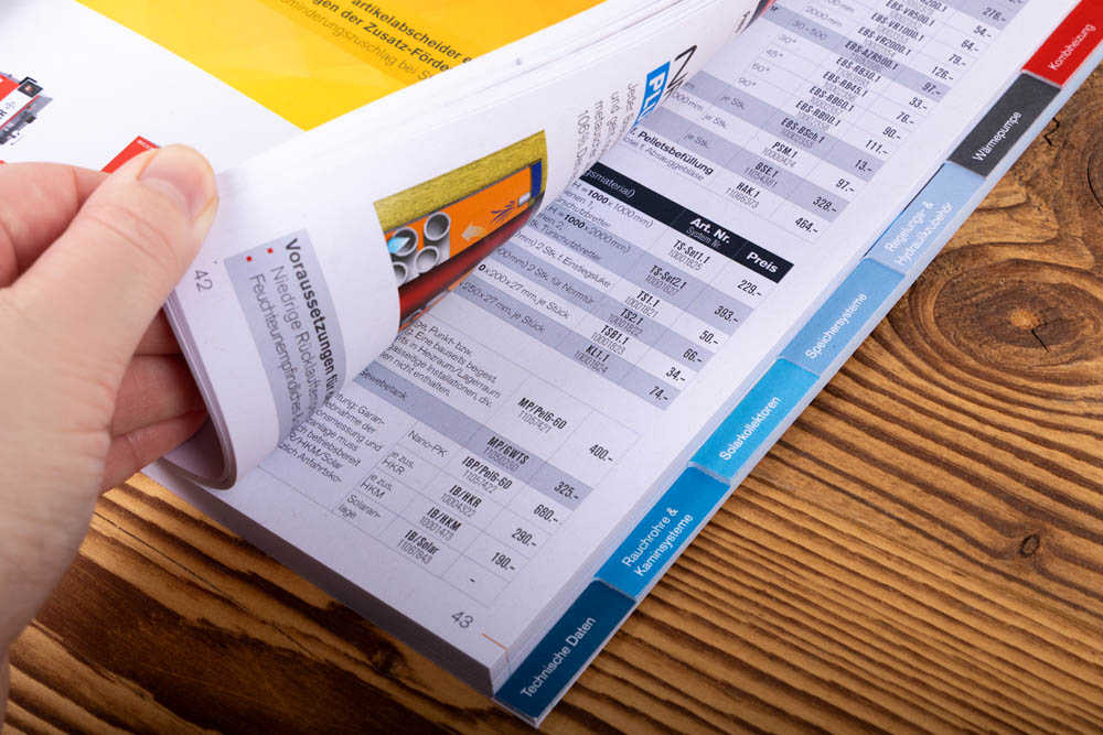 Eine Hand blättert durch die Seiten eines dicken Katalogs oder Verzeichnisses auf einer hölzernen Oberfläche, wobei Spalten mit Zahlen, Produktinformationen und bunten Registerkarten zum Vorschein kommen.