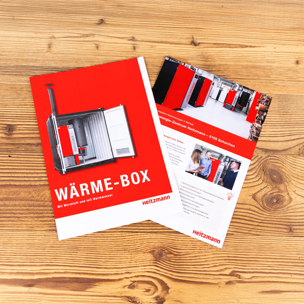 Zwei Heitzmann-Broschüren auf einem Holztisch. Die größere Broschüre zeigt eine rot-weiße Box mit der Aufschrift "WÄRME-BOX" und der Abbildung eines Industrieheizgerätes. Der kleinere Prospekt zeigt mehrere Produktfotos und Text.