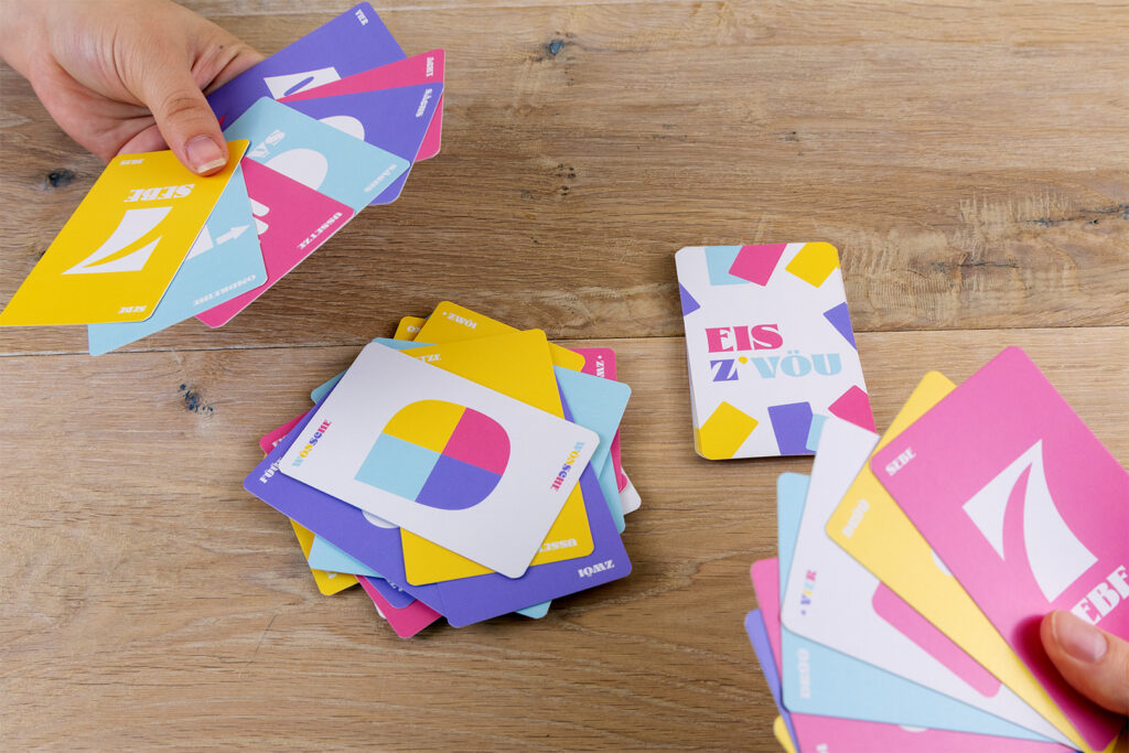 Zwei Personen spielen ein buntes Kartenspiel auf einem Holztisch. Jeder Spieler hält ein Blatt mit Karten mit verschiedenen bunten Formen und Zahlen in der Hand, während weitere Karten auf dem Tisch verteilt sind.