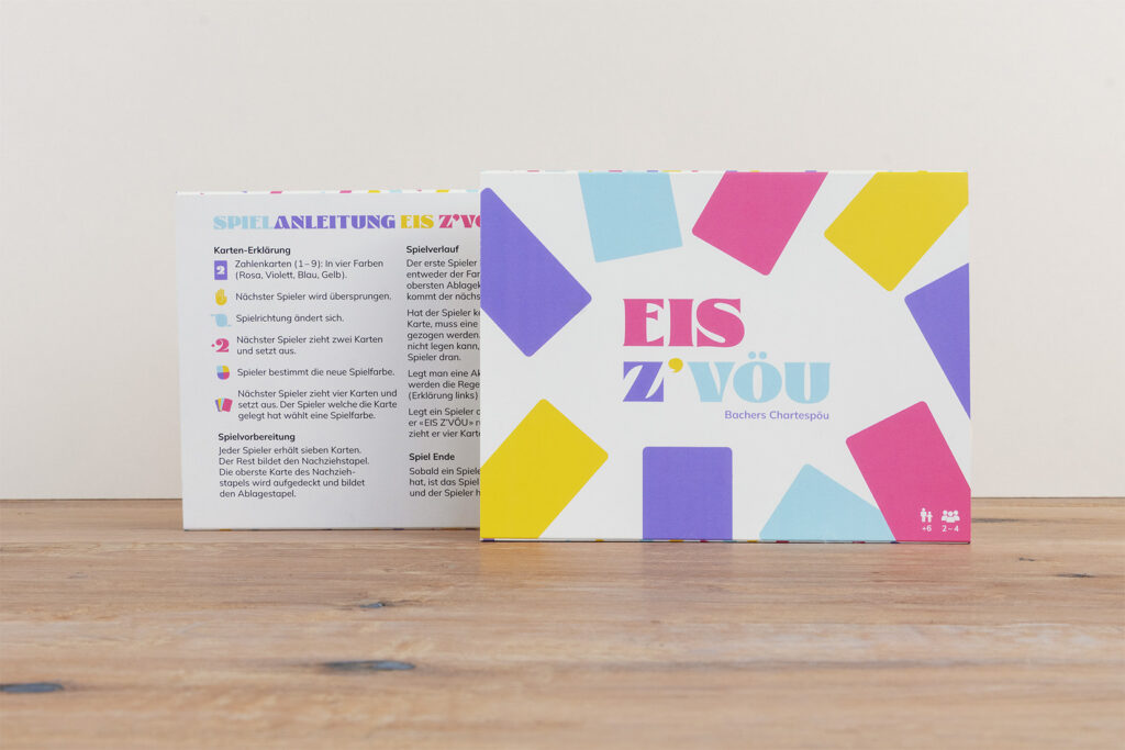 Eine bunte Kartenspielschachtel mit der Aufschrift "EIS Z'VOU" mit bunten Rechtecken auf weißem Hintergrund. Auf der Rückseite der Schachtel ist die Spielanleitung in deutscher Sprache aufgeführt. Die Schachtel steht auf einer Holzfläche.
