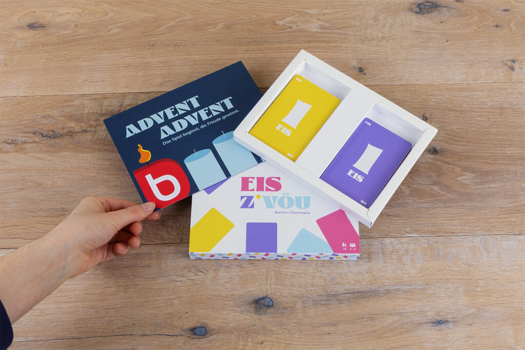 Eine Hand hält eine Karte neben bunten Karten und Schachteln mit den Aufschriften "EIS" und "ADVENT ADVENT" auf einer Holzfläche. Die Verpackung zeigt kräftige Farben, geometrische Formen und deutschen Text.
