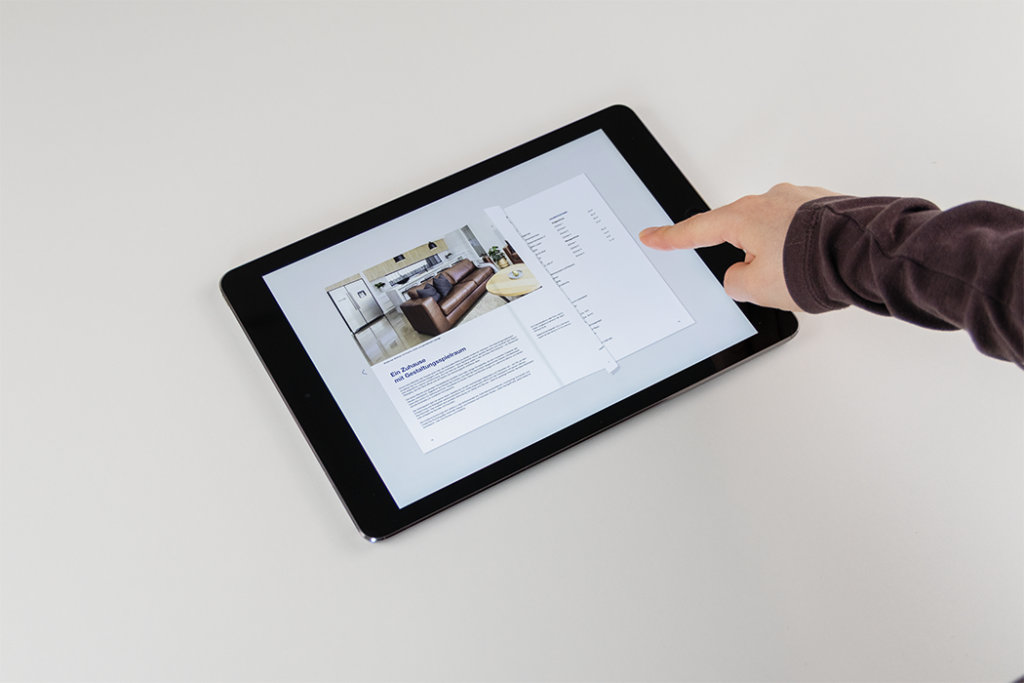 Die Hand einer Person interagiert mit einem Tablet, das eine digitale Zeitschriften- oder Katalogseite mit dem Bild eines modernen Wohnzimmers und etwas Text anzeigt. Das Gerät befindet sich auf einer weißen Oberfläche.