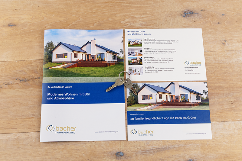 Eine Broschüre, die ein modernes Haus zeigt, liegt aufgeschlagen auf einer hölzernen Oberfläche, auf der ein Paar Hausschlüssel liegt. Die Broschüre zeigt Bilder und Informationen über Immobilien von "bacher immomarketing".