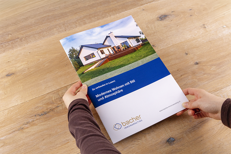 Eine Person hält eine Immobilienbroschüre in der Hand, auf deren Titelseite ein modernes Haus mit einem Rasen abgebildet ist. Die Broschüre hat blaue und weiße Abschnitte mit deutschem Text und dem Logo der "bacher IMMOBILIEN AG" am unteren Rand.