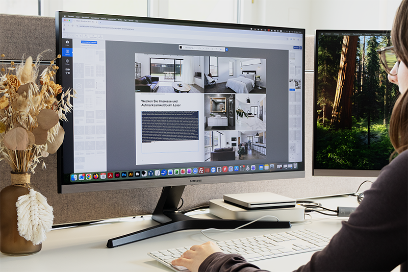 Eine Person arbeitet an einem Schreibtisch mit einem großen Monitor, auf dem ein Design-Layout mit Bildern und Text angezeigt wird. Ein weiterer Monitor zeigt eine Waldszene. Auf dem Schreibtisch befinden sich Trockenblumen in einer Vase und Büromaterialien.
