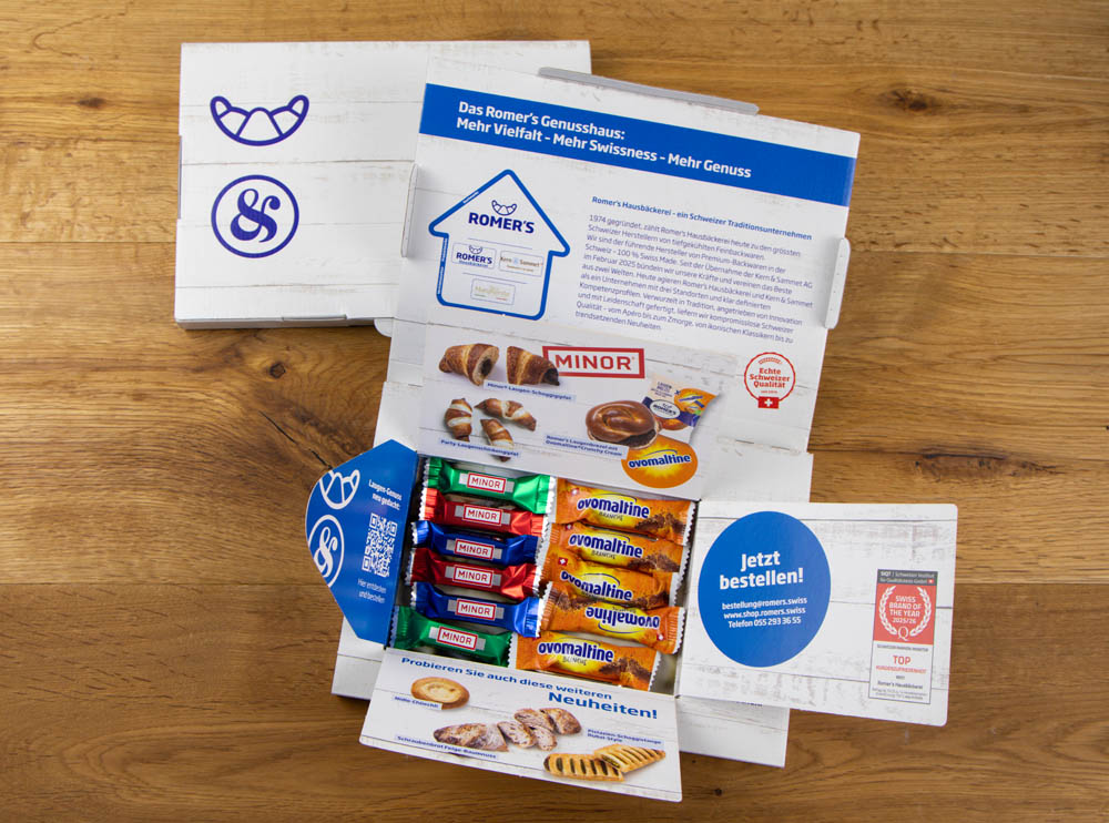 Eine geöffnete Musterbox auf einem Holztisch zeigt sechs Minor-Schokoladentafeln und fünf Ovomaltine-Schokoladentafeln, umgeben von Produktbildern, Informationen und der Aufschrift Jetzt bestellen! (Bestellen Sie jetzt!) in deutscher Sprache.