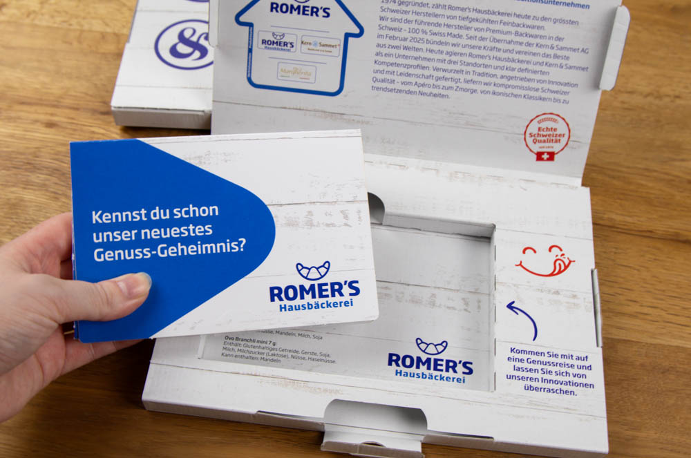 Eine Hand hält eine blau-weiße Schachtel mit der Aufschrift "NEU! gleich reinschauen" und "ROMER'S Hausbäckerei" über einen offenen Pizzakarton auf einem Holztisch. Die Verpackung ist mit einer einfachen, verspielten Smiley-Grafik versehen.