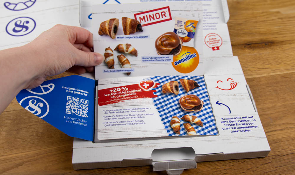 Eine Hand hebt die Klappe einer Produktschachtel und enthüllt Bilder und Beschreibungen von croissantförmigen Snacks, die mit Ovomaltine-Creme gefüllt sind. Auf der Verpackung sind die Marke MINOR und zusätzliche Werbeinformationen abgebildet.