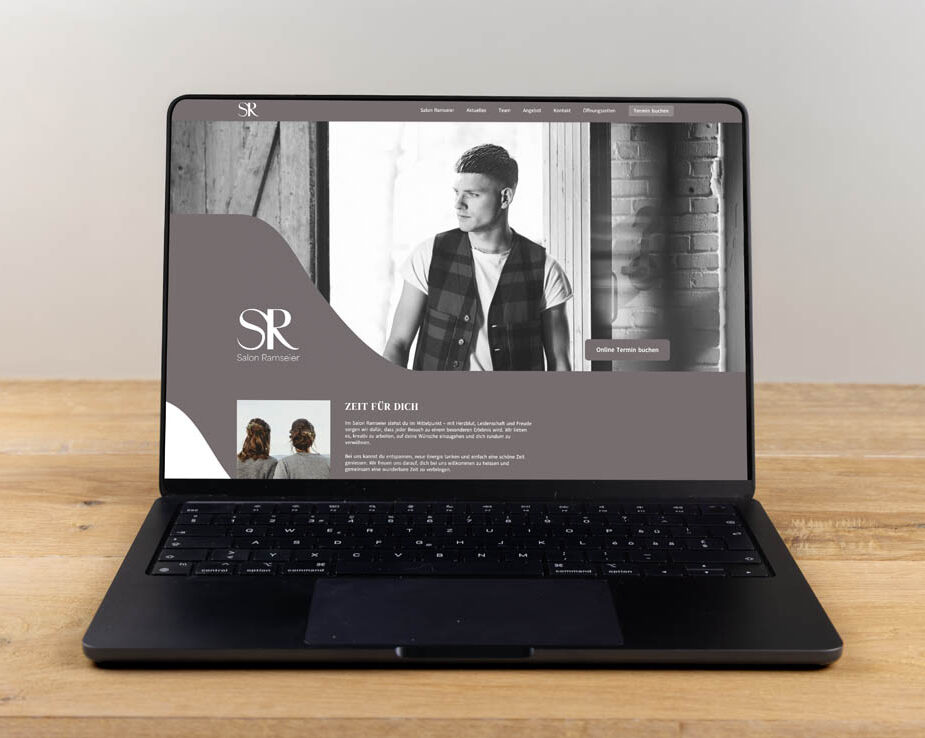 Ein Laptop auf einem Holztisch zeigt eine Website für den "Salon Ramsdorf" mit dem Schwarz-Weiß-Foto eines Mannes mit gestyltem Haar, Text und Navigationslinks auf dem Bildschirm.