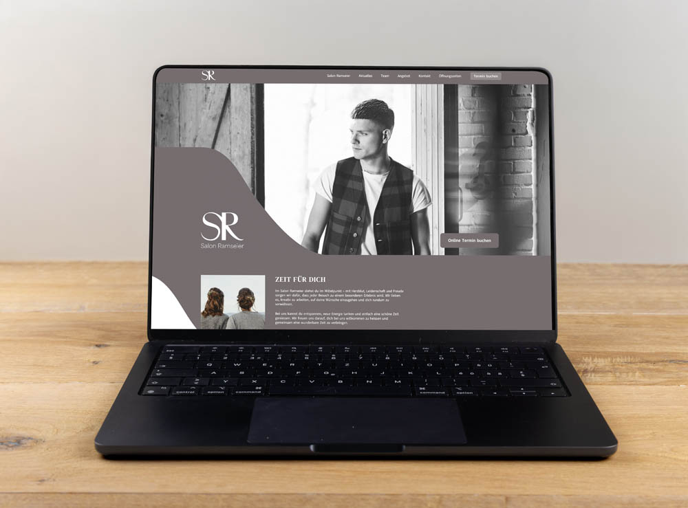 Ein Laptop auf einem Holztisch zeigt eine Website für den "Salon Ramsdorf" mit dem Schwarz-Weiß-Foto eines Mannes mit gestyltem Haar, Text und Navigationslinks auf dem Bildschirm.