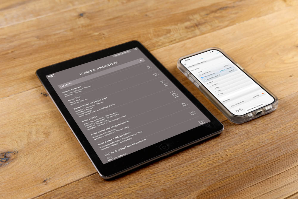 Ein Tablet und ein Smartphone liegen auf einer Holzoberfläche. Das Tablet zeigt ein Menü auf Deutsch an, während das Smartphone eine Kalender- oder Terminplan-App anzeigt. Beide Geräte sind schwarz und eingeschaltet.