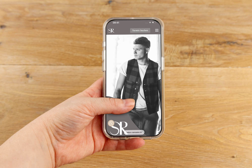 Eine Hand hält ein Smartphone, auf dem ein Schwarz-Weiß-Foto einer Person in einer ärmellosen karierten Weste und einem weißen Hemd mit Holzhintergrund zu sehen ist. Auf dem Bildschirm erscheint eine Buchungsoption.