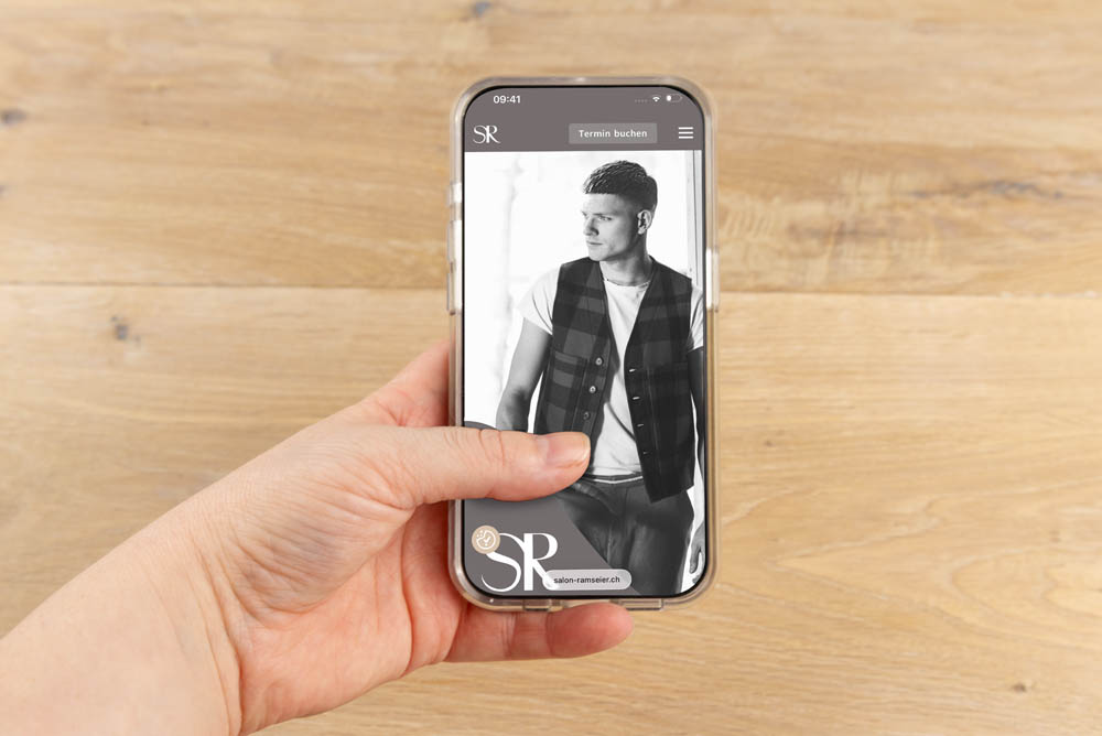 Eine Hand hält ein Smartphone, auf dem ein Schwarz-Weiß-Foto eines Mannes in karierter Weste und T-Shirt zu sehen ist. Auf dem Bildschirm des Telefons sind außerdem ein Logo und Menüsymbole zu sehen. Der Hintergrund ist eine helle Holzoberfläche.