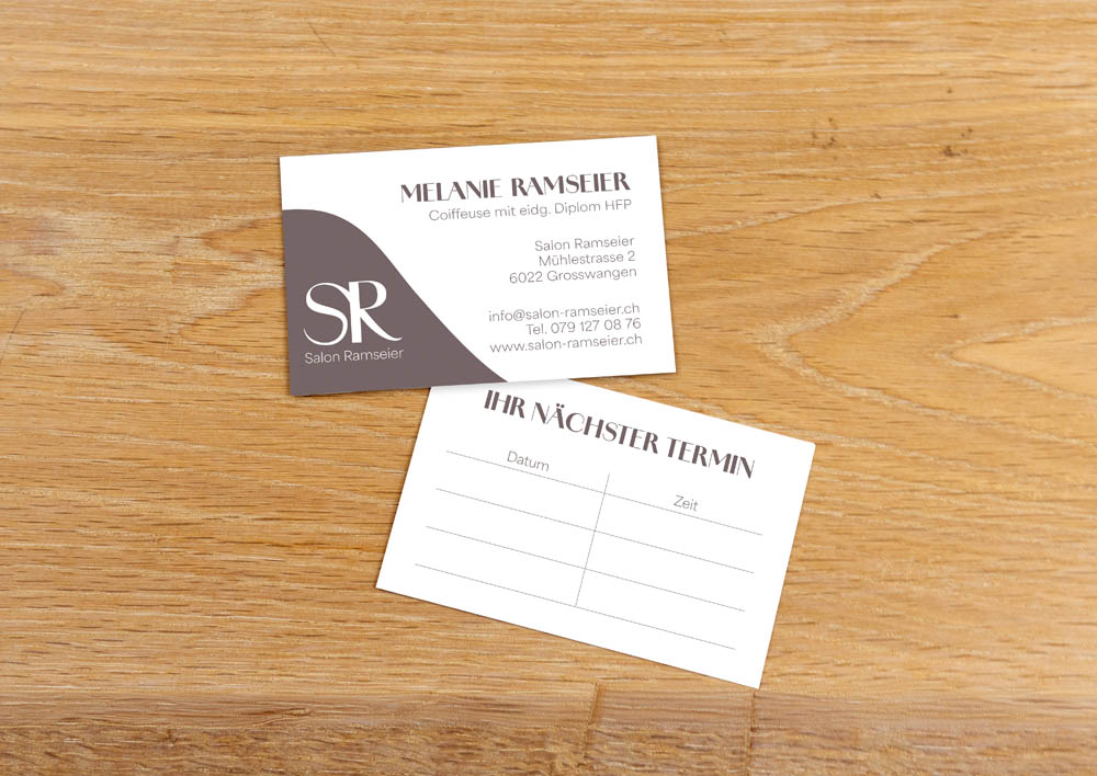 Zwei Visitenkarten auf einer Holzoberfläche. Eine Karte zeigt die Kontaktdaten des Salons mit einem Logo und dem Namen "Melanie Ramseier", die andere Karte ist eine Terminkarte mit Feldern für Datum und Uhrzeit.