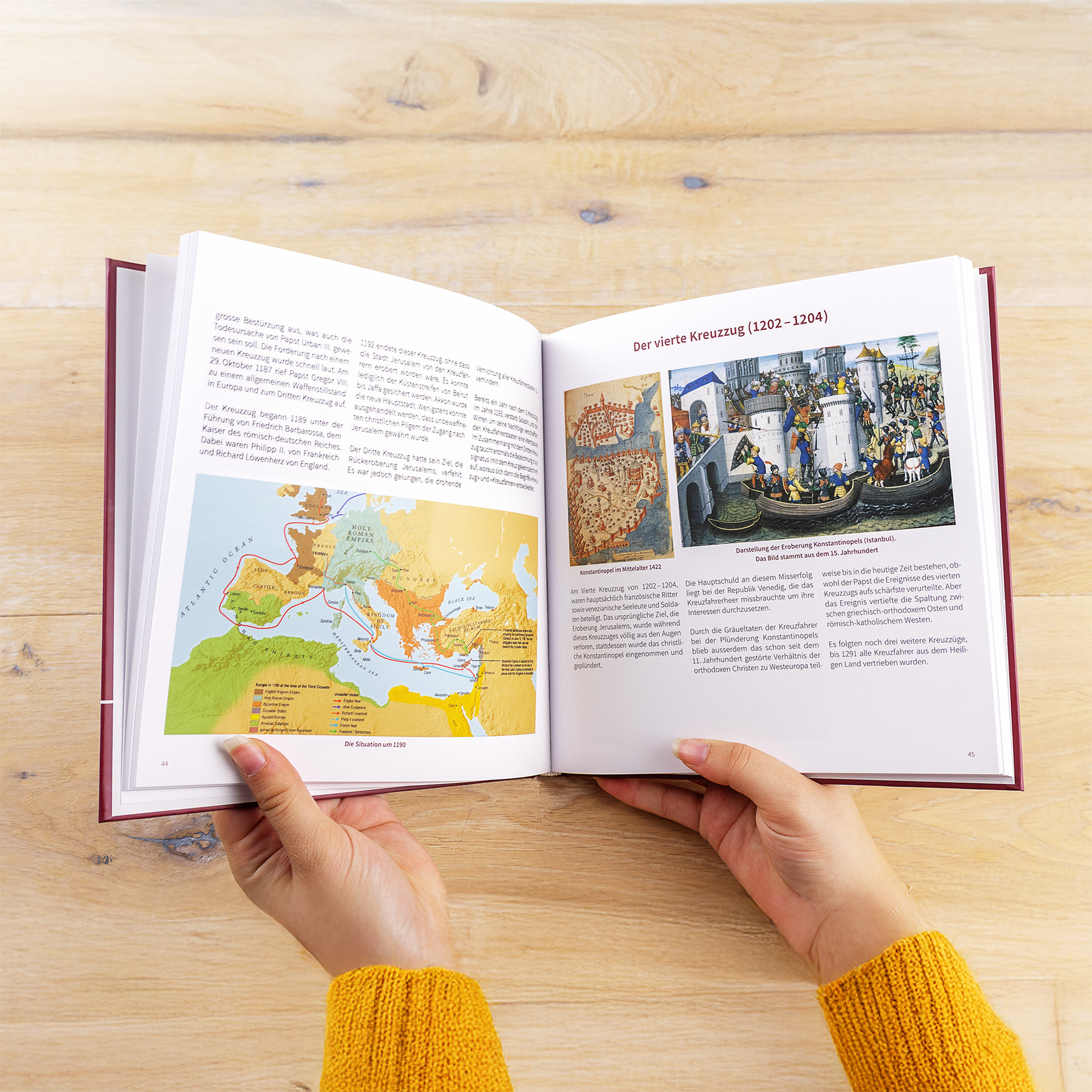 Eine Person in einem gelben Pullover hält ein Buch auf einem hellen Holztisch aufgeschlagen, das Seiten mit deutschem Text, eine Europakarte und eine Abbildung des Vierten Kreuzzugs (1202-1204) zeigt.