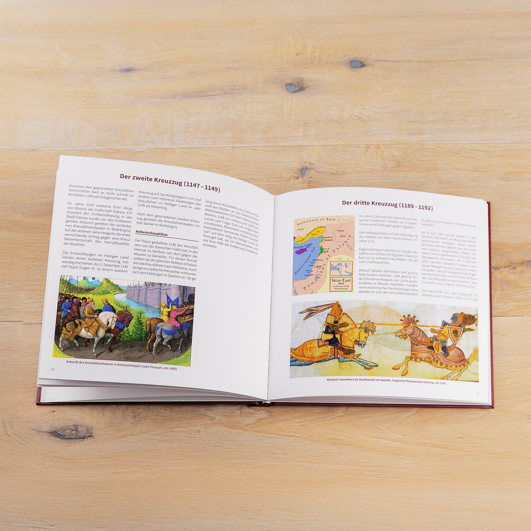 Ein aufgeschlagenes Buch auf einer hölzernen Oberfläche zeigt einen deutschen Text mit farbenfrohen Illustrationen von mittelalterlichen Rittern und einer Karte, die den zweiten und dritten Kreuzzug beschreiben.