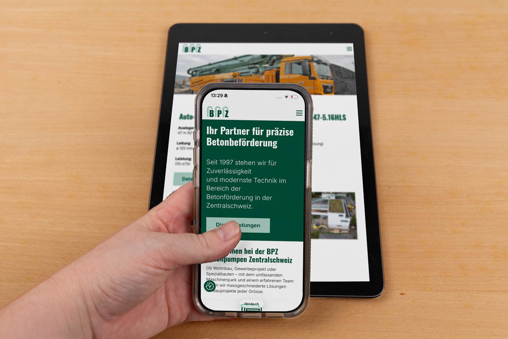 Eine Hand hält ein Smartphone, auf dem die Website eines Betontransportunternehmens angezeigt wird, während ein Tablet, auf dem dieselbe Website angezeigt wird, auf einem Holztisch im Hintergrund liegt.