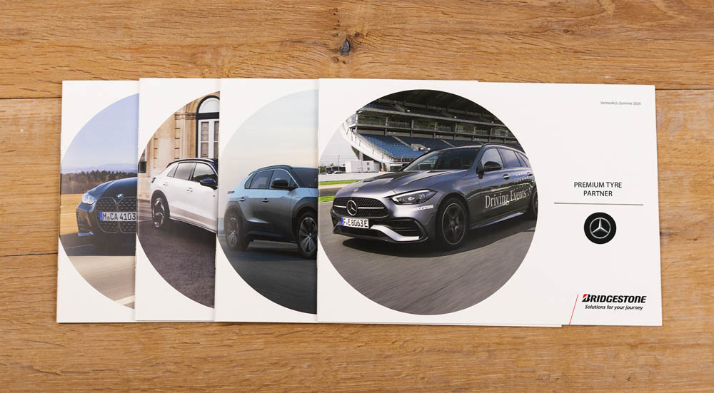 Vier sich überlappende Karten mit verschiedenen Mercedes-Benz Fahrzeugen, die auf einer Holzunterlage platziert sind. Die Karte ganz rechts zeigt ein Mercedes-Auto und die Logos von Mercedes-Benz und Bridgestone.