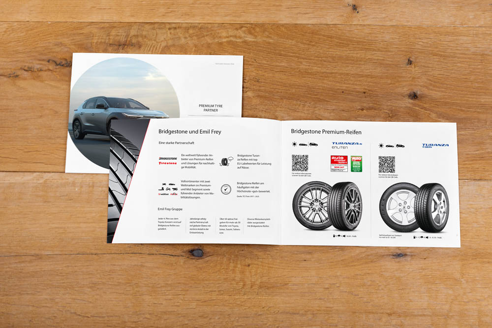 Eine geöffnete Autobroschüre auf einer Holzoberfläche zeigt Bilder von Autoreifen, einem Auto und Reifenspezifikationen in deutscher Sprache. Die Broschüre enthält QR-Codes und verschiedene Symbole zu den Eigenschaften und der Leistung von Reifen.