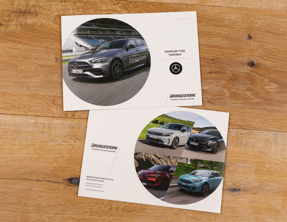 Zwei Postkarten auf einer Holzunterlage, jeweils mit Abbildungen von Autos und dem Bridgestone-Logo: oben ein silberner Mercedes-Benz, unten drei Autos.