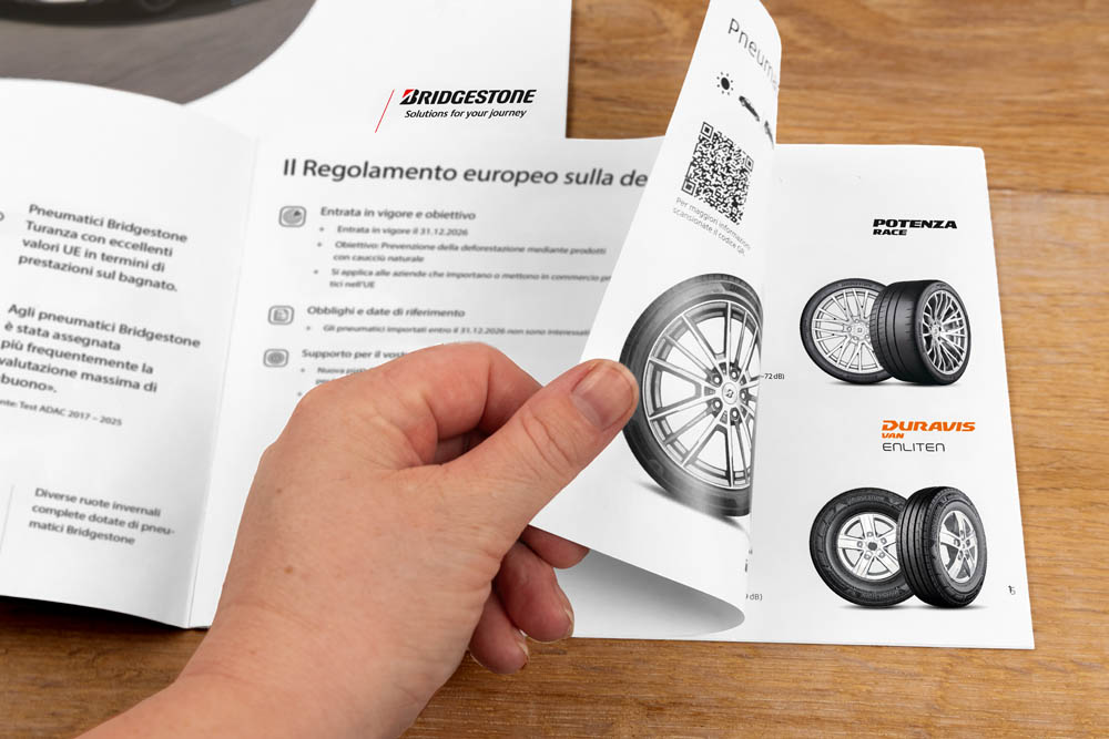 Eine Hand blättert durch eine Bridgestone-Reifenbroschüre auf einer hölzernen Unterlage, die Potenza Race- und Duravis Enliten-Reifen, QR-Codes und italienischen Text zeigt.