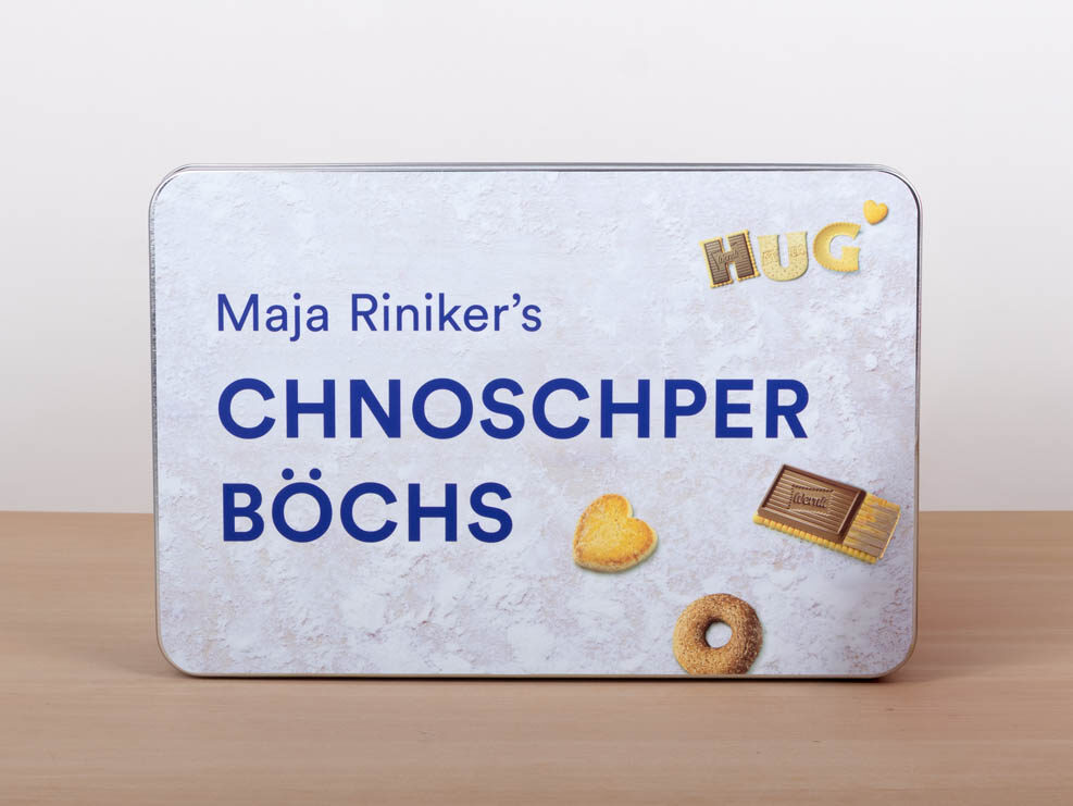 Eine Dose mit der Aufschrift "Maja Riniker's CHNOSCHPER BOECHS", auf der Kekse abgebildet sind, steht neben einer durchsichtigen, rot verpackten Packung mit Keksen auf einer Holzfläche.