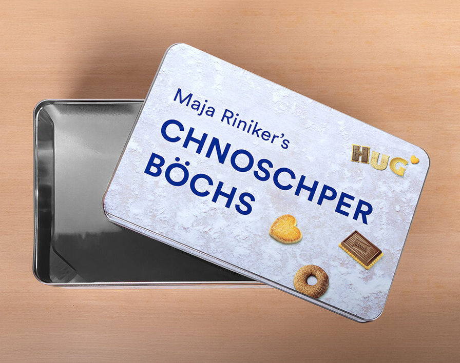Eine rechteckige Dose mit der Aufschrift "Maja Riniker's Chnoschper Boechs" steht leicht geöffnet da und gibt den Blick auf rot verpackte Kekse frei. Auf dem Deckel der Dose sind drei Biscuits und das HUG-Logo auf hellem Grund abgebildet.
