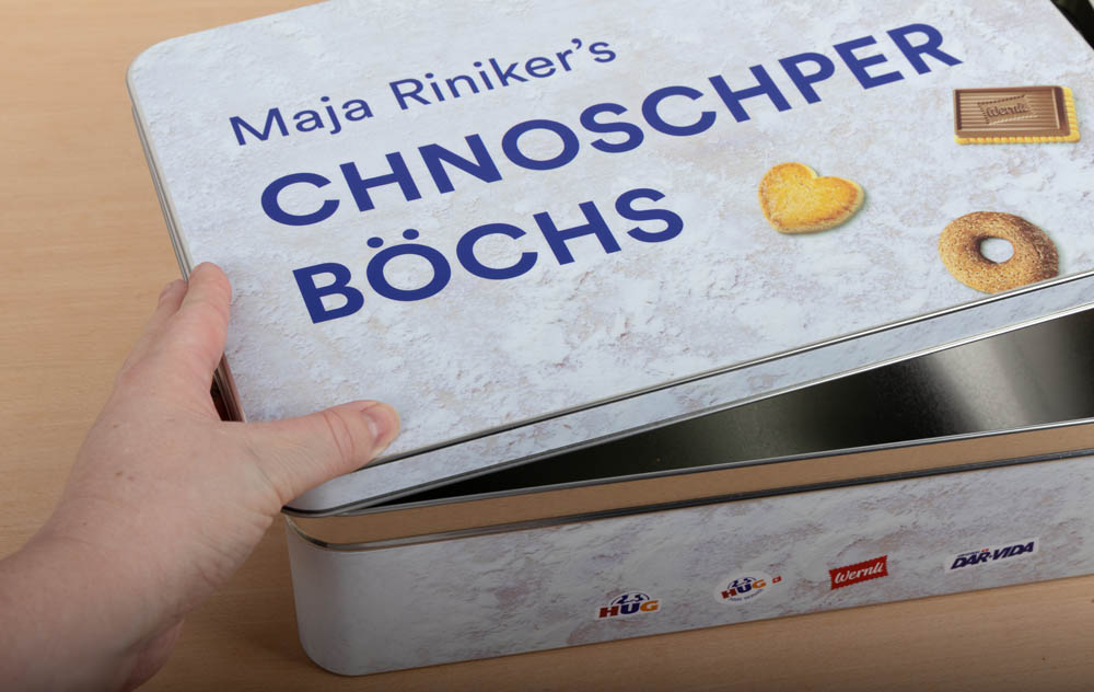 Eine Hand öffnet eine Metalldose mit der Aufschrift "Maja Riniker's Chnoschper Boechs" und Abbildungen von Keksen und Snacks auf dem Deckel, die auf einer hellen Holzfläche steht.