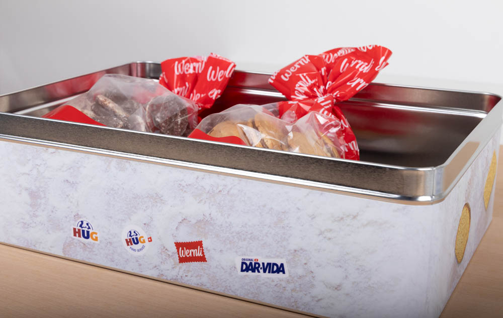 Eine Metalldose, die durchsichtige Tüten mit verschiedenen Keksen enthält, die mit einer rot-weißen Schleife verschnürt sind. Auf der Seite der Dose sind Aufkleber mit den Logos "HUG", "Wernli" und "DAR-VIDA" angebracht.