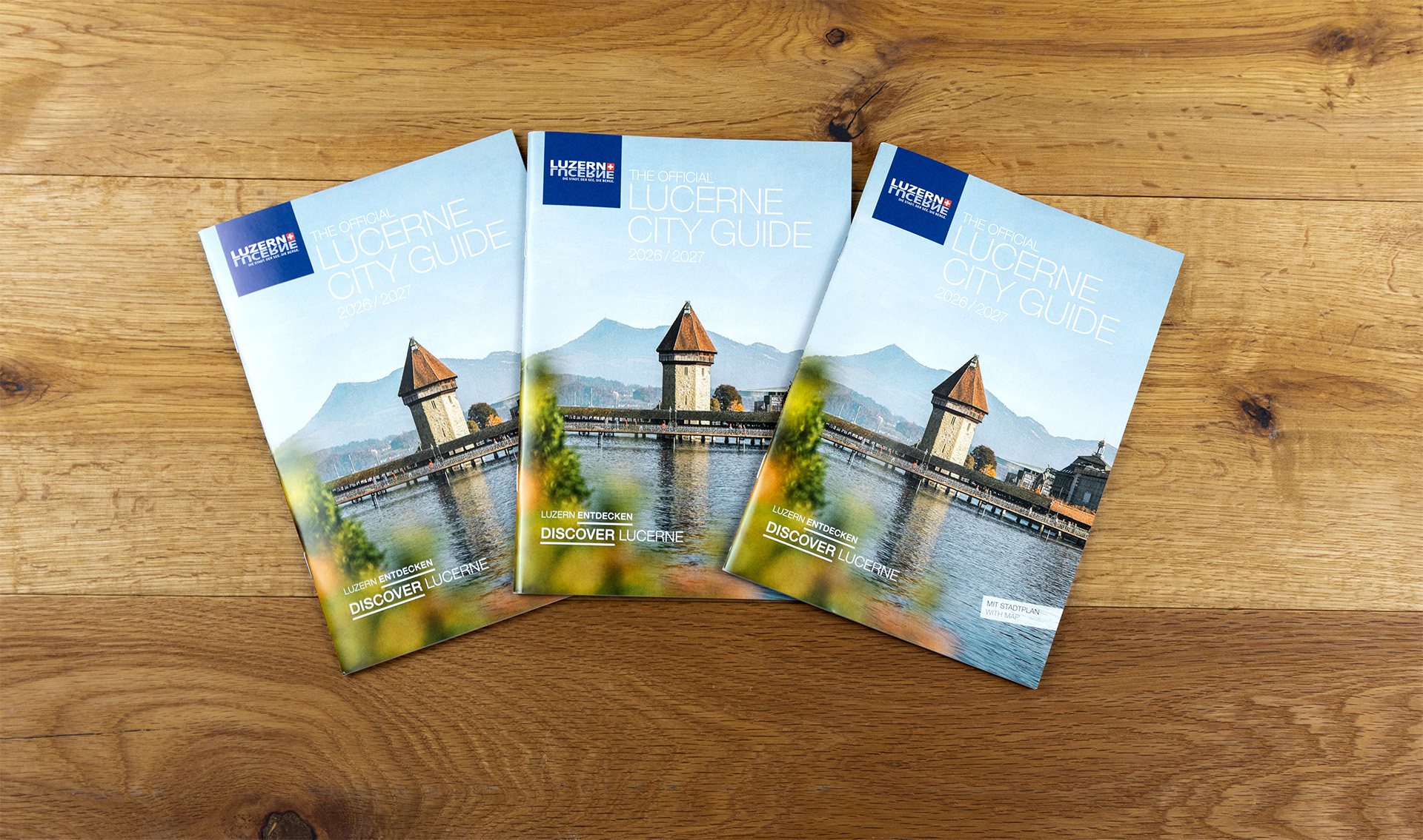 Drei Exemplare des "Stadtführers Luzern" auf Holzunterlage, Umschlag zeigt Kapellbrücke und Wasserturm mit Alpen und blauem Himmel.