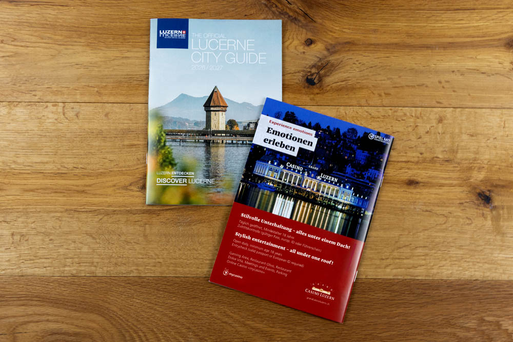 Zwei Broschüren liegen auf einer Holzplatte. Die obere ist der offizielle Stadtführer von Luzern und zeigt die Berge und einen historischen Turm am Wasser. Die untere Broschüre, rot mit deutschem Text und nächtlichen Gebäudebildern, enthält ein Bridgestone-Logo.