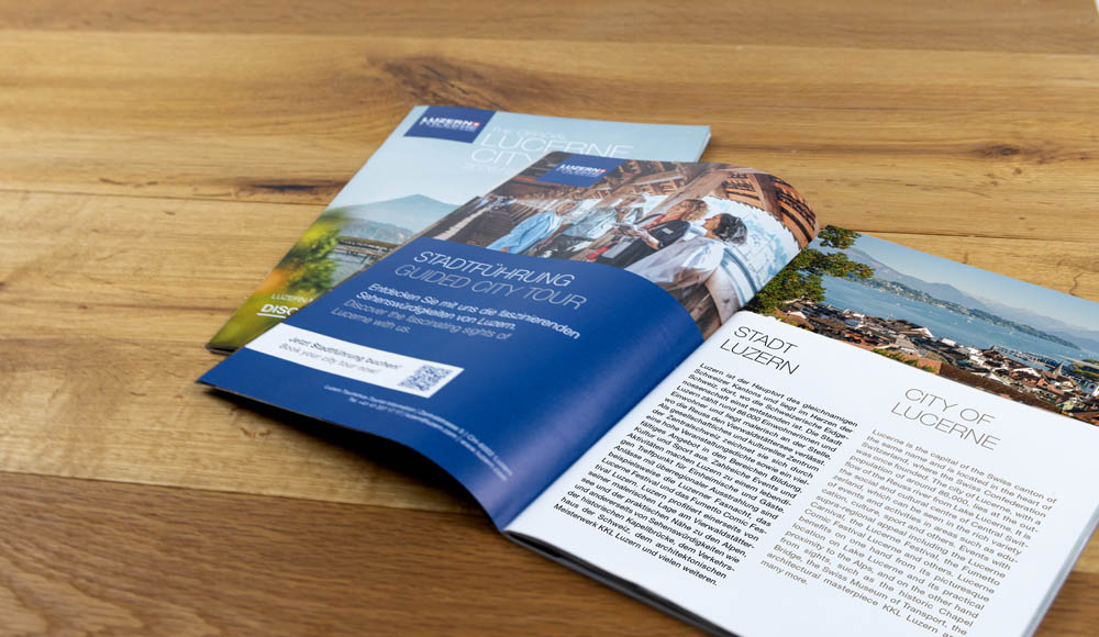 Zwei aufgeschlagene Broschüren auf einem Holztisch zeigen Informationen und Bilder über die Stadt Luzern, darunter eine von Bridgestone gesponserte Stadtführungsseite, eine Stadtbeschreibung und Landschaftsfotos.