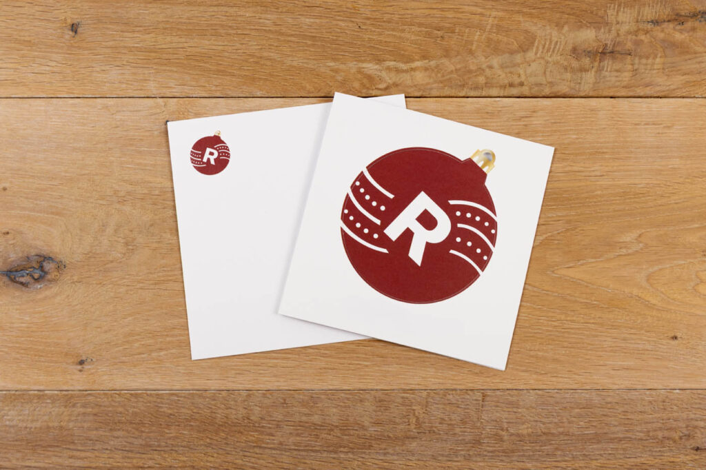Zwei weiße quadratische Karten auf einer hölzernen Oberfläche; die obere Karte zeigt ein großes rotes Weihnachtsornament mit einem weißen "R" und dekorativen Linien, während die Karte darunter eine kleinere Version desselben Ornaments zeigt.