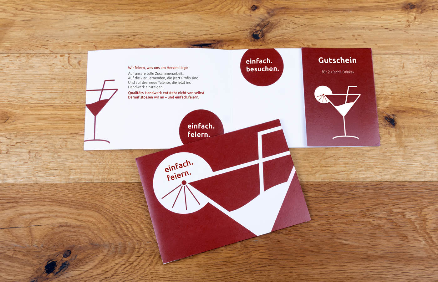Ein rot-weißer Geschenkgutschein mit minimalistischen Cocktailglas-Illustrationen ist auf einer Holzoberfläche abgebildet. Der deutsche Text beinhaltet "einfach. feiern." und "Gutschein für 2x40€ Drinks.".