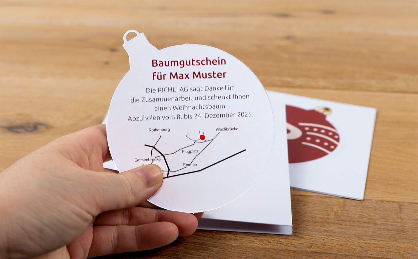 Eine Hand hält einen Gutschein in Form einer Weihnachtskugel mit einem deutschen Text, der einen kostenlosen Weihnachtsbaum verspricht, einschließlich einer Karte. Im Hintergrund liegt ein weißer Umschlag mit der Abbildung einer roten Weihnachtskugel auf einem Holztisch.