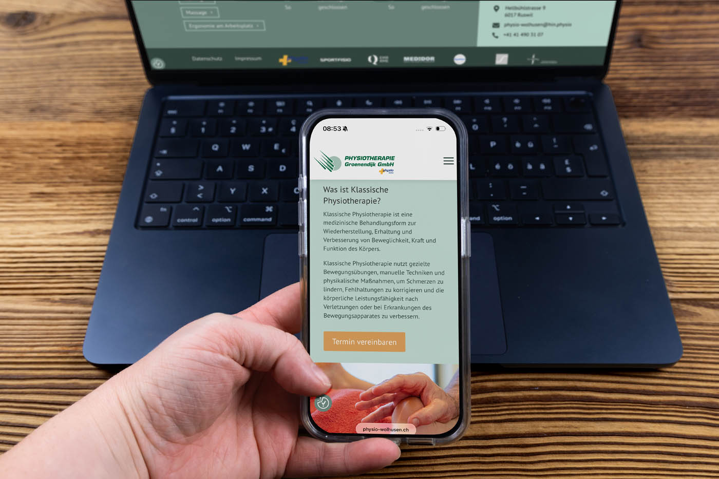 Eine Person hält ein Smartphone, auf dem die deutsche Website von Physio-Wolhusen zu sehen ist, vor einen geöffneten Laptop auf einem Holztisch. Auf dem Bildschirm des Smartphones sind Informationen zur klassischen Physiotherapie zu sehen.