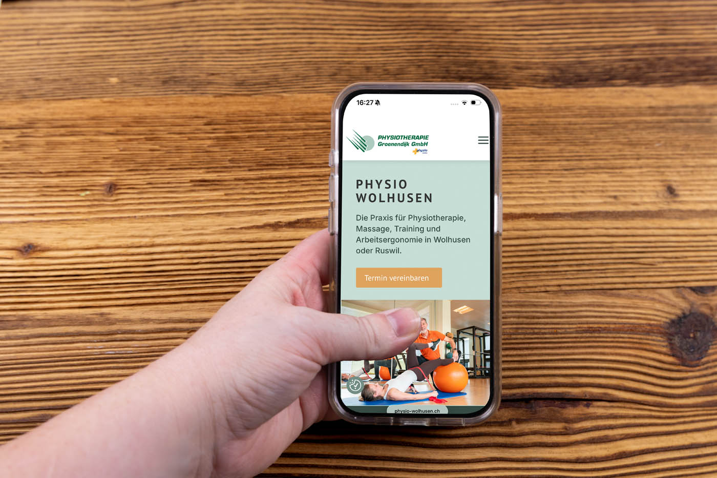 Eine Hand, die ein Smartphone hält, zeigt die Website von Physio-Wolhusen, die physiotherapeutische Dienstleistungen anbietet; das Smartphone liegt auf einer Holzunterlage.
