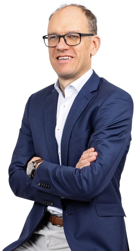 Ein Mann mit Brille, blauem Sakko, weißem Hemd und einer Armbanduhr lächelt mit verschränkten Armen vor einem schlichten weißen Hintergrund und strahlt Selbstvertrauen und die Bereitschaft aus, erfolgreiche Projekte zu leiten.