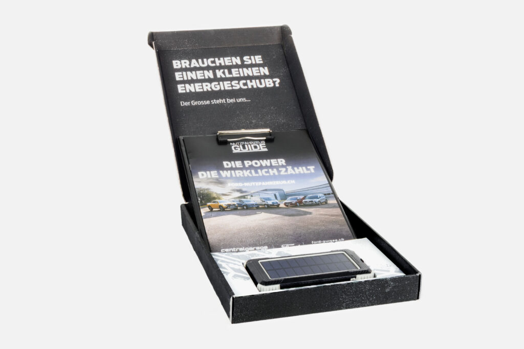Eine geöffnete schwarze Schachtel, in der vor einem schlichten weißen Hintergrund ein Heft mit deutschem Text und ein kleines solarbetriebenes Gerät zu sehen sind.