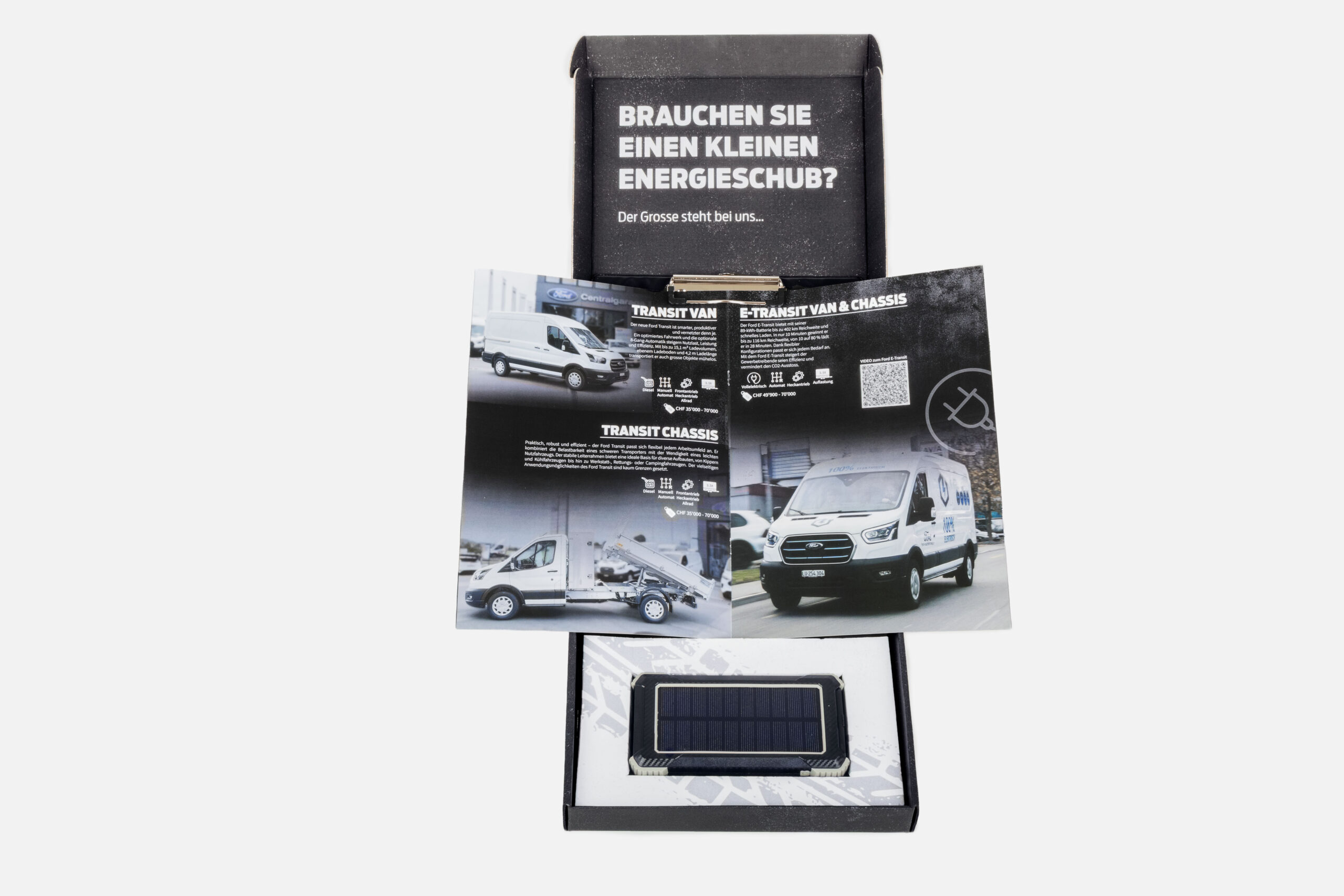 Offene Schachtel mit Broschüren zum Ford Transit und einem rechteckigen Gegenstand mit einem Solarpanel; Text in deutscher Sprache auf dem Deckel der Schachtel.