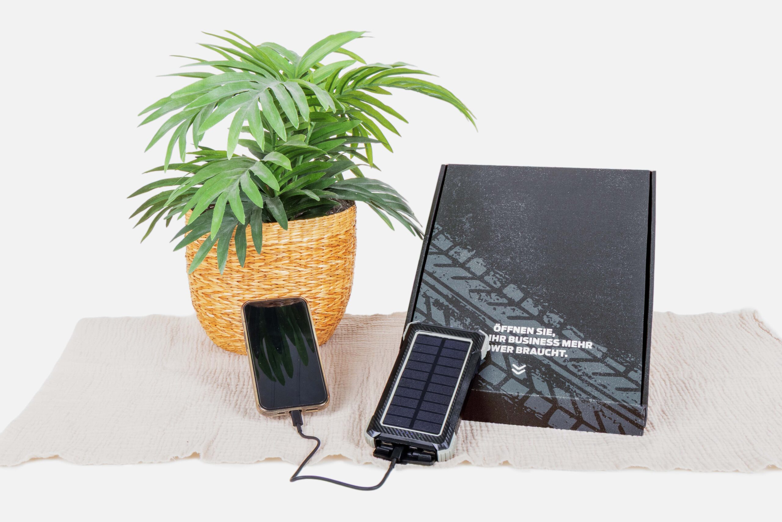 Eine Topfpflanze, ein Smartphone, das über ein Solarladegerät aufgeladen wird, und eine schwarze Schachtel mit Reifenprofil-Motiv und deutscher Beschriftung sind auf einem hellen Tuch vor weißem Hintergrund angeordnet.