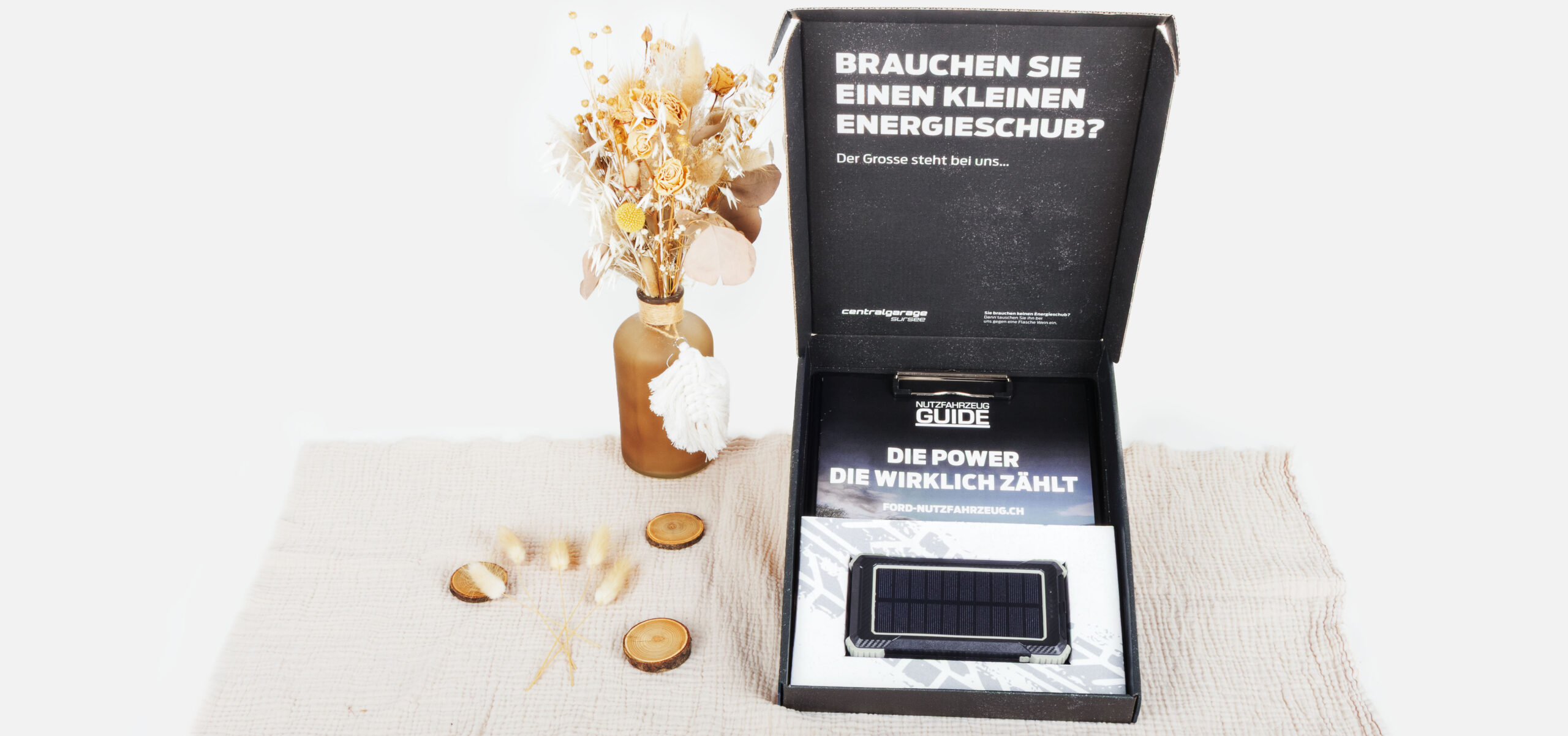 Eine Geschenkbox mit einer Solar-Powerbank, Broschüren und getrockneten Blumen in einer Vase auf einem beigen Tuch. Auf der Box steht auf Deutsch: „Brauchen Sie einen kleinen Energieschub? Die Power, die sich wirklich lohnt.“.