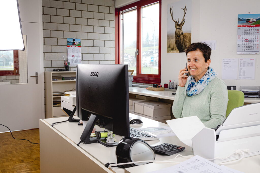 Eine Frau sitzt an einem Schreibtisch in einem Büro und lächelt, während sie telefoniert. Sie sitzt vor einem Computerbildschirm, um sie herum liegen Papiere, ein Drucker und Büromaterial. An der Wand hängt ein großes Foto eines Rehs.