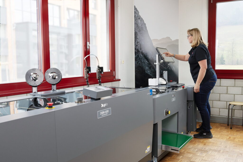 Eine Frau bedient eine große Duplo-DGM-420-Maschine in einem hellen Raum mit roten Fensterrahmen und Blick auf die Berge.