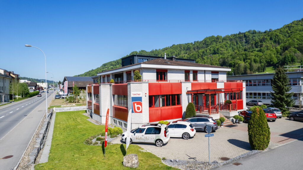 Ein modernes Gebäude mit roten Akzenten, mehreren geparkten Autos und einem grünen Rasen steht an einer Straße, im Hintergrund sind Hügel und Bäume unter einem strahlend blauen Himmel zu sehen.