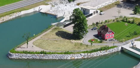 Luftaufnahme eines kleinen, dreieckigen Grundstücks an einem Fluss mit einem roten Haus, einem Baum und einer eingezäunten Rasenfläche, die einen nachhaltigen Ansatz bei der Landnutzung verdeutlicht. Zu sehen sind außerdem eine Straße und eine Brücke, die von blaugrünem Wasser und Betonkonstruktionen umgeben sind.